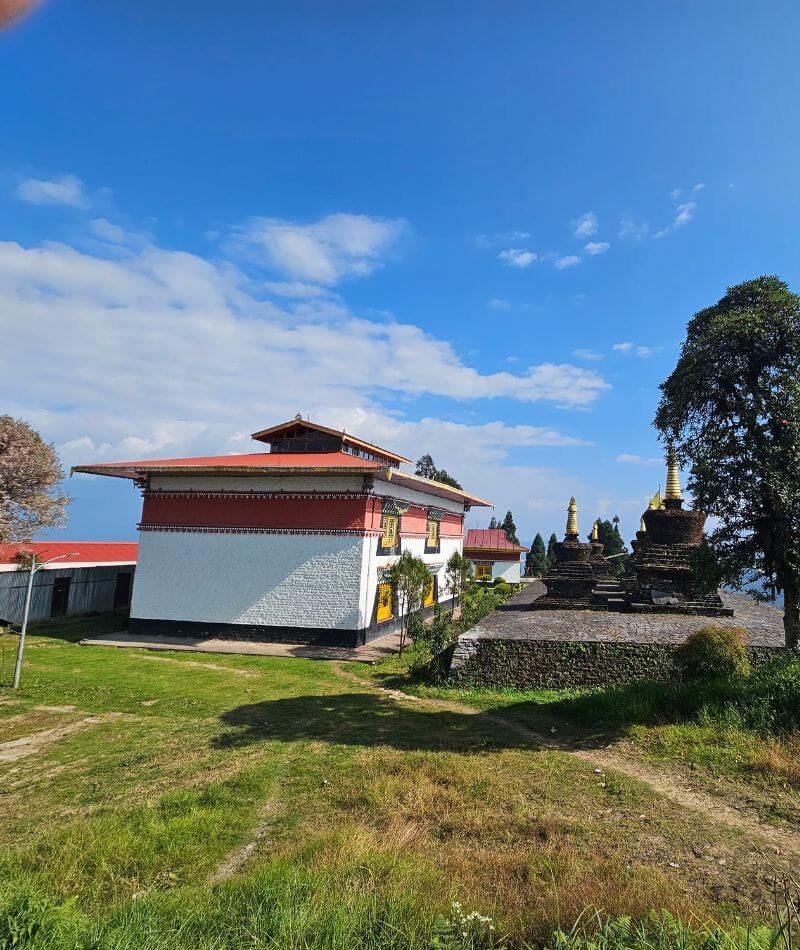 A side-facing view of a traditional Tibetan Buddhist temple with white walls, a red tiered roof, and yellow-trimmed windows. The building is surrounded by a green lawn with manicured bushes and a row of dark, bell-shaped stupas with golden spires on the left. A tall, thin flagpole stands in the foreground on a sunny day under a clear blue sky.