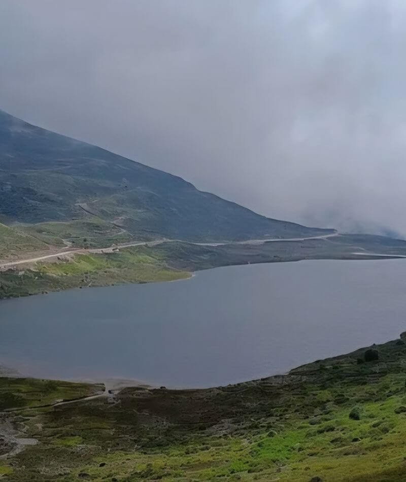 A high-altitude, wide-angle shot of a large, still lake surrounded by barren, grassy hills. A narrow, winding mountain road traces the edge of the water and climbs up the steep slope of a green mountain on the left. The scene is shrouded in heavy mist and low-hanging gray clouds, creating a somber and isolated atmosphere.