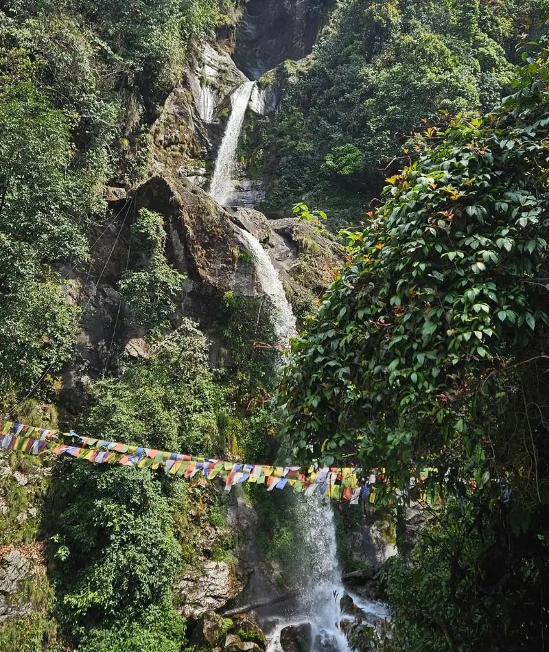 A vertical, eye-level shot of a tall, slender waterfall cascading down a dark, rocky cliff face. The water falls in two distinct tiers, surrounded by dense, vibrant green moss and tropical foliage. In the lower foreground, a long string of colorful Buddhist prayer flags (blue, white, red, green, and yellow) is strung horizontally across the view. The scene is bright, with sunlight illuminating the falling water and the surrounding leaves.