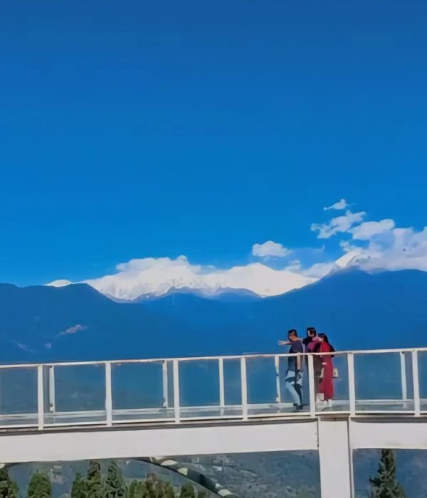 A wide shot featuring three people standing on a high-altitude glass skywalk or bridge with white railings. They are looking out toward a breathtaking mountain range. In the distance, massive, snow-covered peaks rise above a layer of dark blue mountain ridges. The sky above is a vibrant, solid blue with a few wispy white clouds clinging to the mountain summits.