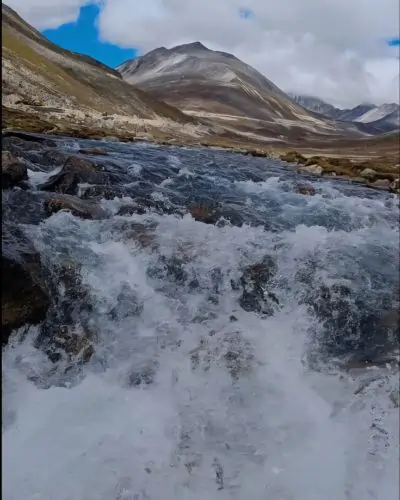 A fast-flowing mountain stream with white rapids rushing over dark rocks in the foreground. In the background, a large, barren mountain peak rises toward a bright blue sky filled with large, white clouds.