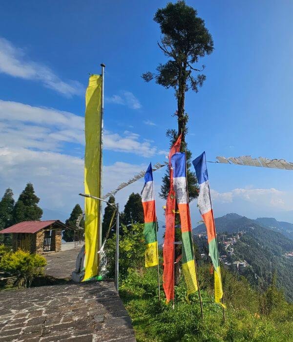 A paved outdoor area on a mountain ridge featuring a small white building with a red roof. Several tall, colorful prayer flags stand along the edge of the slope, overlooking a deep valley and distant, forested hills under a bright blue sky with scattered clouds.